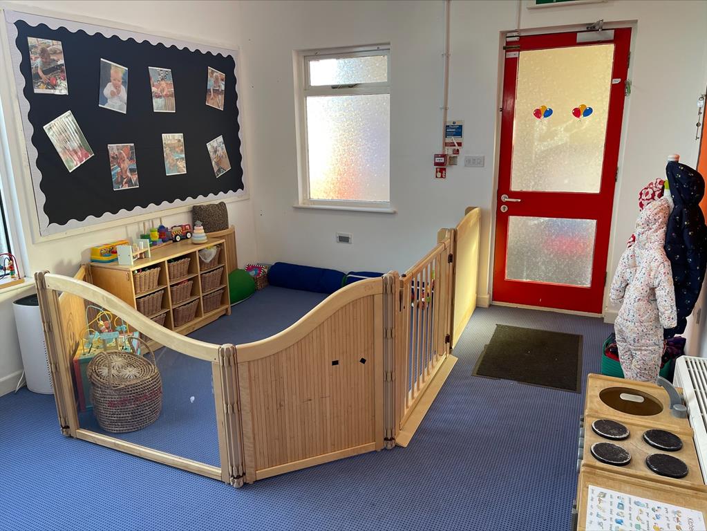 Nursery room with wooden play pen area, toy cooker and kitchen area, photos of babies and toddlers playing on a display board. Coats hanging up.